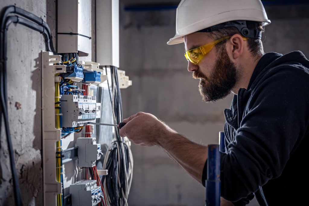 A male electrician works in a switchboard with an electrical connecting cable, connects the equipment with tools.